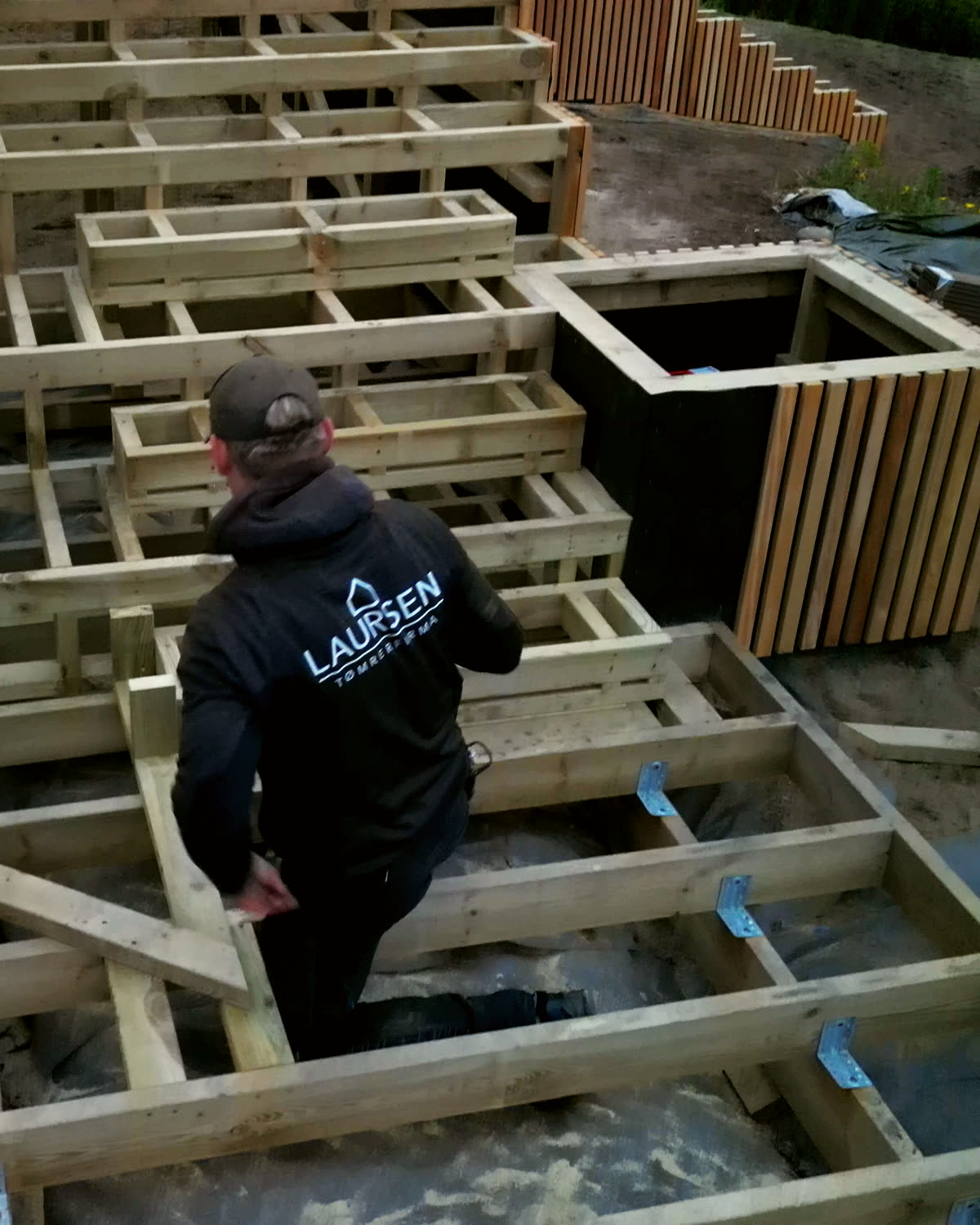 Tømrer i Laursen-trøje går gennem terrassens bjælkelag ved en plantekasse med lameller